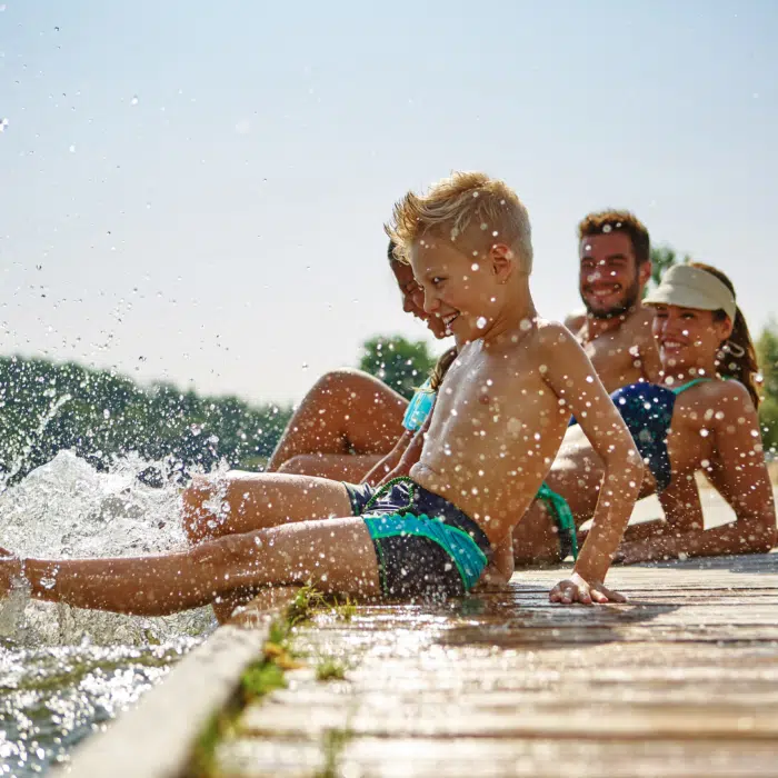 Ihr seht eine Familie am Steg eines Sees sitzen. JUFA Hotels bietet tollen Sommerurlaub an schönen Seen für die ganze Familie.