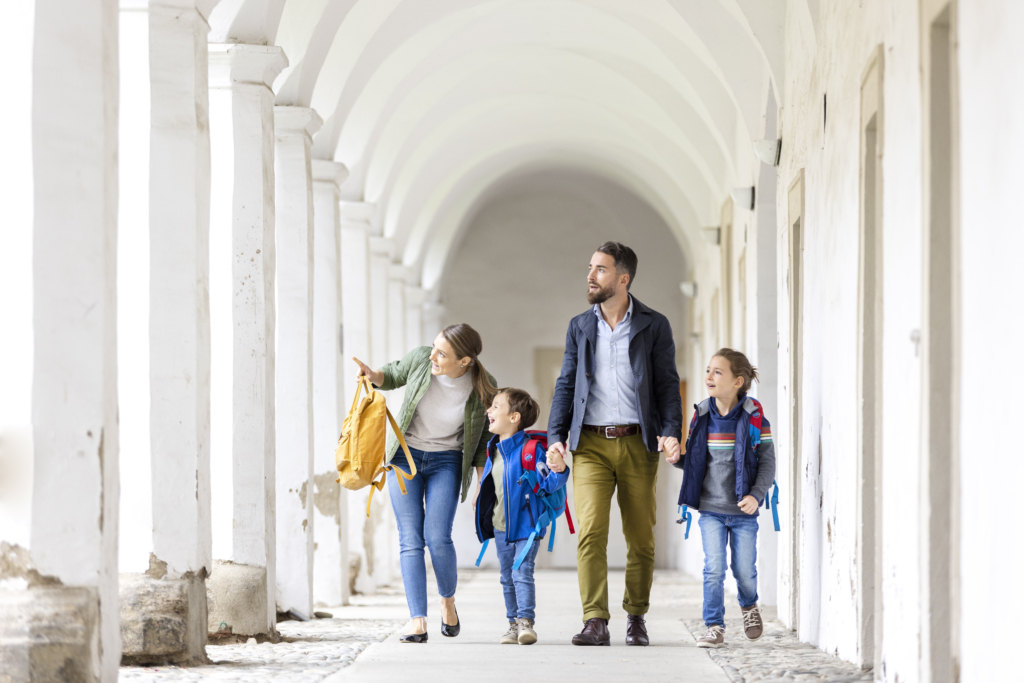 Ihr seht eine Familie von vorne mit Muter, Vater, Tochter, Sohn. Die Mutter zeigt auf etwas und alle Blicke folgen nach links. Die Familie spaziert durch die Klosterbögen vom JUFA Hotel Judenburg. Der Ort für erholsamen Familienurlaub und einen unvergesslichen Winter- und Wanderurlaub.