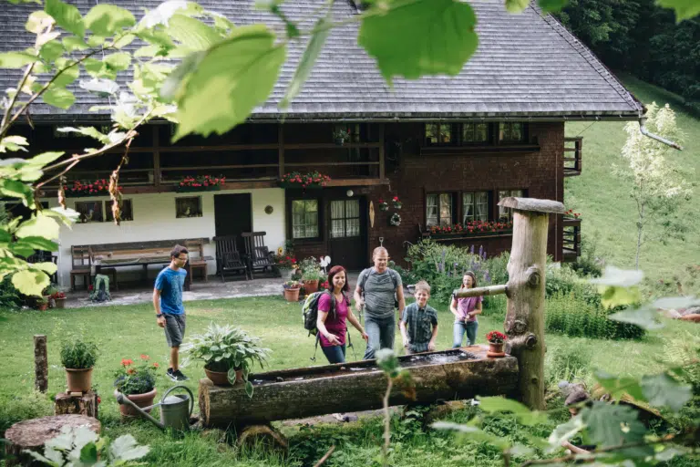Ihr seht eine Familie, die sich auf einer Alm an einem Brunnen erfrischt und abkühlt. JUFA Hotels bietet Ihnen den Ort für erlebnisreichen Natururlaub für die ganze Familie.