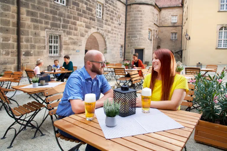 Ihr seht ein Paar am Tisch sitzend im Innenhof des JUFA Hotel Festung Rosenberg Kronach. Der Ort für kinderfreundlichen und erlebnisreichen Urlaub für die ganze Familie.