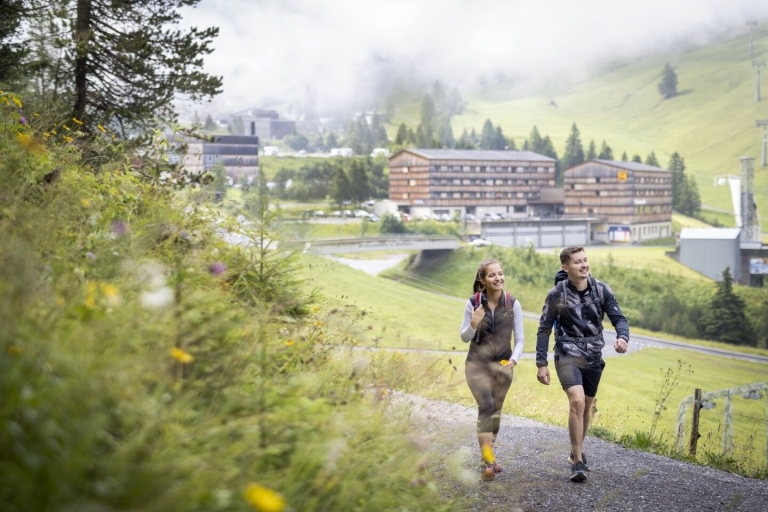 Ihr seht das JUFA Hotel Malbun mit zwei motivierten Wanderern im Vordergrund
