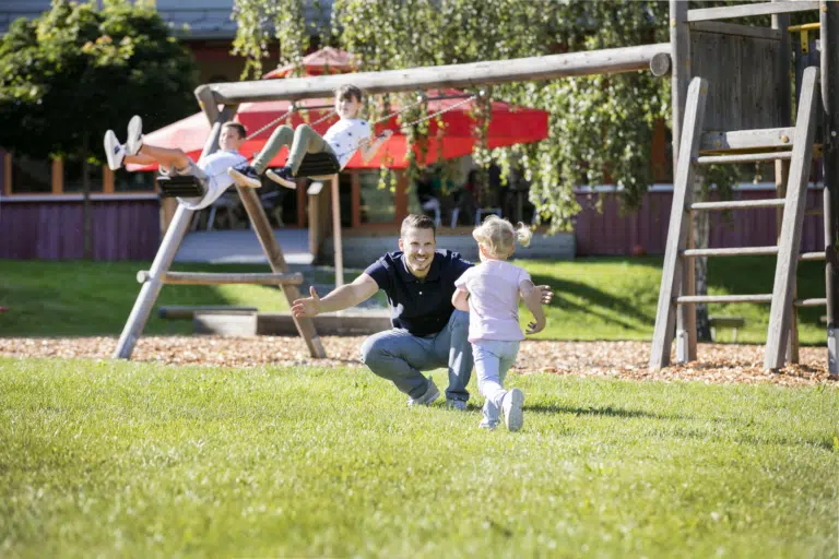 Ihr seht eine Familie spielen und schaukeln am Spielplatz des JUFA Hotels Waldviertel. Der Ort für kinderfreundlichen und erlebnisreichen Urlaub für die ganze Familie.