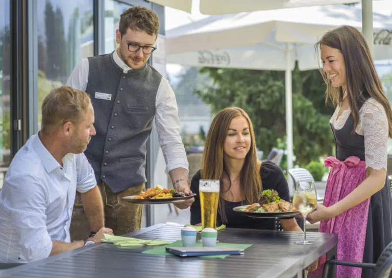 Ihr seht ein Paar und einen Kellner sowie eine Kellnerin auf der Terrasse im JUFA Hotel Altenmarkt***.