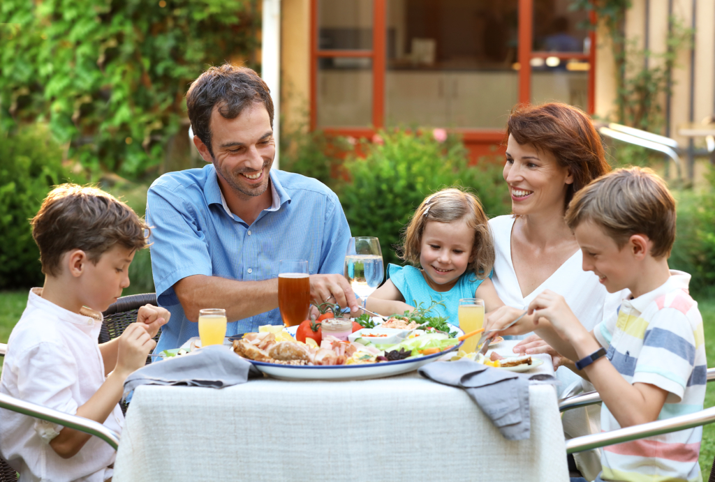 Sie sehen eine Familie bei einer Jause auf der Terrasse des JUFA Hotels Pöllau - Bio-Landerlebnis. Der Ort für erholsamen Familienurlaub und einen unvergesslichen Winter- und Wanderurlaub.
