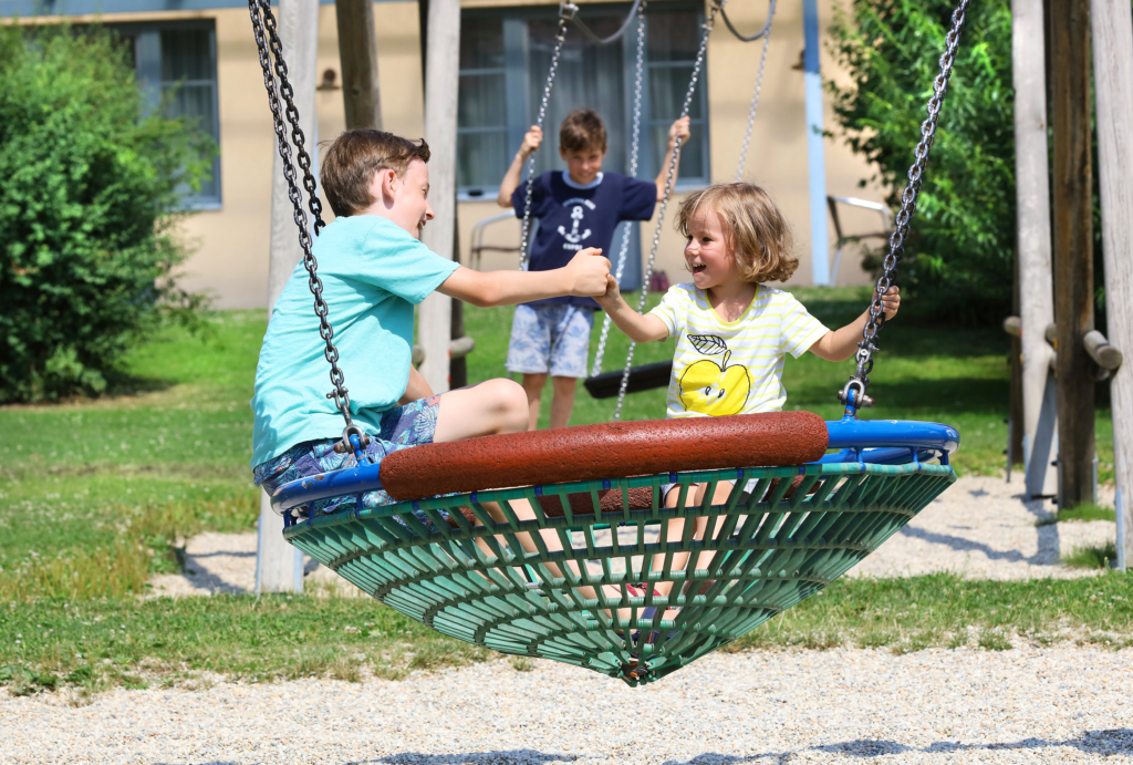 Sie sehen Kinder auf einer Korbschaukel am Spielplatz des JUFA Hotels Pöllau - Bio-Landerlebnis. Der Ort für erholsamen Familienurlaub und einen unvergesslichen Winter- und Wanderurlaub.