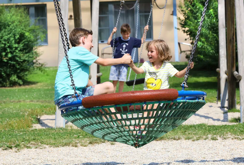 Sie sehen Kinder auf einer Korbschaukel am Spielplatz des JUFA Hotels Pöllau - Bio-Landerlebnis. Der Ort für erholsamen Familienurlaub und einen unvergesslichen Winter- und Wanderurlaub.