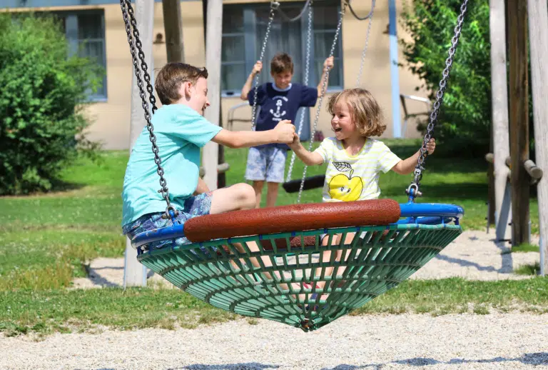 Sie sehen Kinder auf einer Korbschaukel am Spielplatz des JUFA Hotels Pöllau - Bio-Landerlebnis. Der Ort für erholsamen Familienurlaub und einen unvergesslichen Winter- und Wanderurlaub.