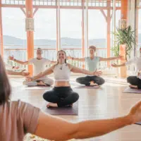 Ihr seht Menschen im Schneidersitz mit ausgebreiteten Armen bei einer Yoga-Atemübung.