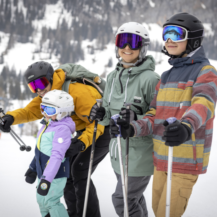 Ihr seht eine Familie mit Vater, Tochter, Mutter und Sohn mit Skihelmen und Skibrillen auf Ski in verschneiter Winterlandschaft.