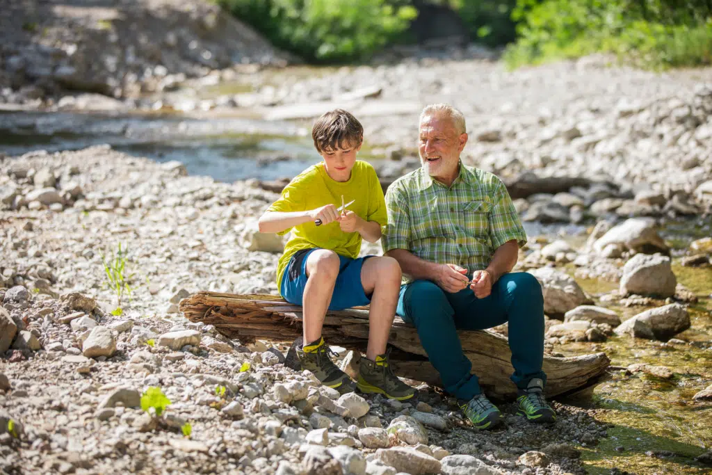 Ihr seht einen Großvater mit seinen Enkel sitzend am Wasser beim Stecken schnitzen. JUFA Hotels bietet erholsamen Familienurlaub und einen unvergesslichen Winter- und Wanderurlaub.
