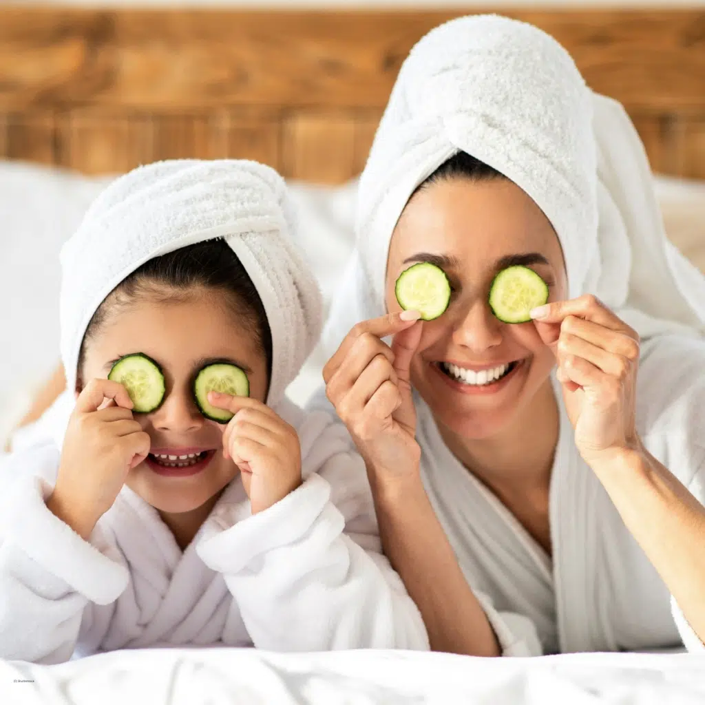Mutter und Tochter liegen in weißen Bademänteln mit Handtuchturban auf dem Bett und halten sich Gurkenscheiben vor die Augen bei einer Wellness-Rauszeit.