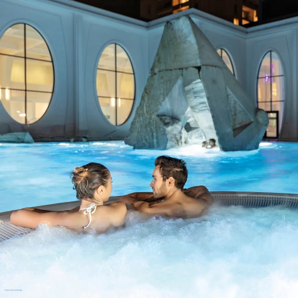 Paar entspannt abends im spudelnden Whirlpool eines Thermenbereichs mit Pyramiden-Skulptur bei einem herbstlichen Wellnessurlaub.