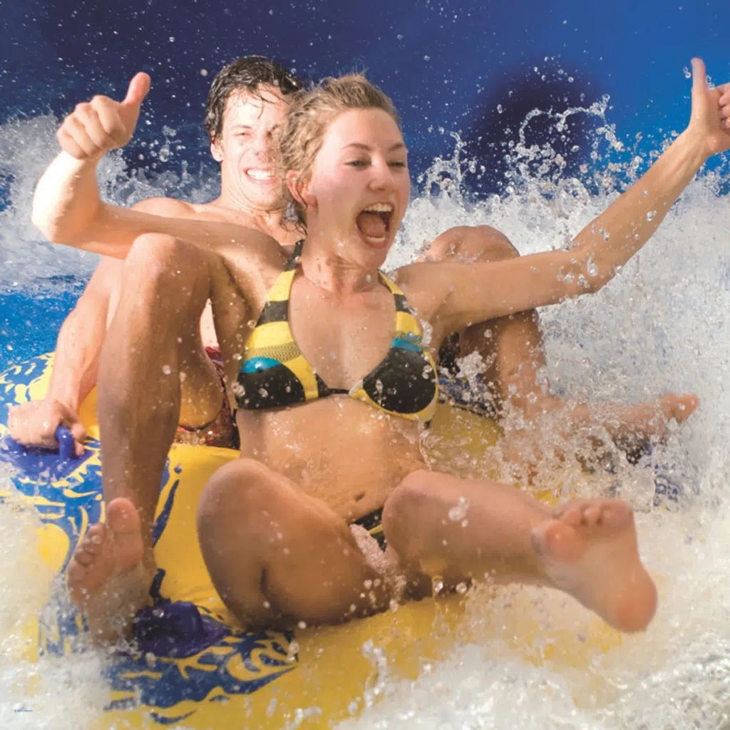 Junge Frau und Mann rutschen lachend auf einem gelben Reifen durch die Wasserrutsche in einem Erlebnisbad.
