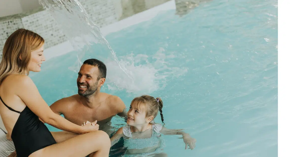 Mutter sitzt am Beckenrand, während Vater und Tochter unter einem Wasserfall im Hallenbad spielen.