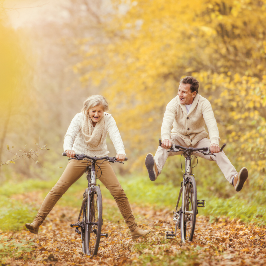 Älteres Paar fährt lachend und mit ausgestreckten Beinen auf Fahrrädern über einen mit Herbstlaub bedeckten Weg.