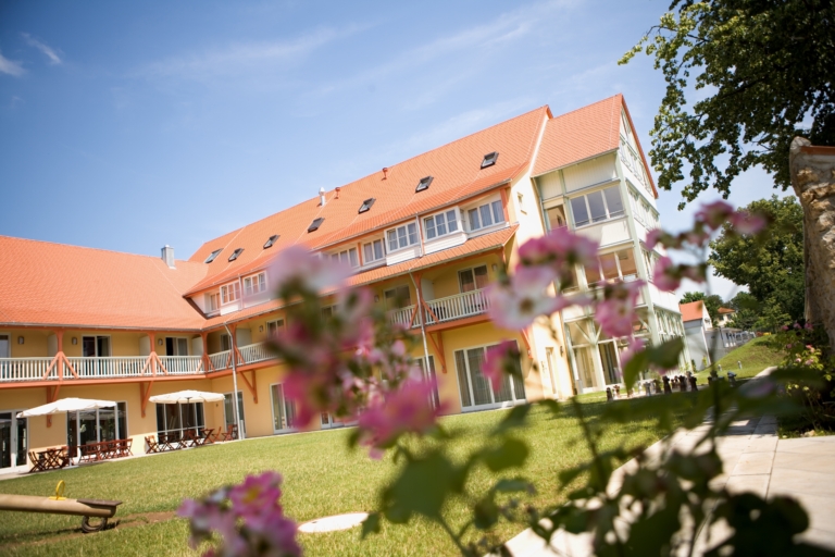 Außenansicht des JUFA Hotel Nördlingen mit gelber Fassade, rotem Ziegeldach, Balkonen und grüner Liegewiese im Sommer.