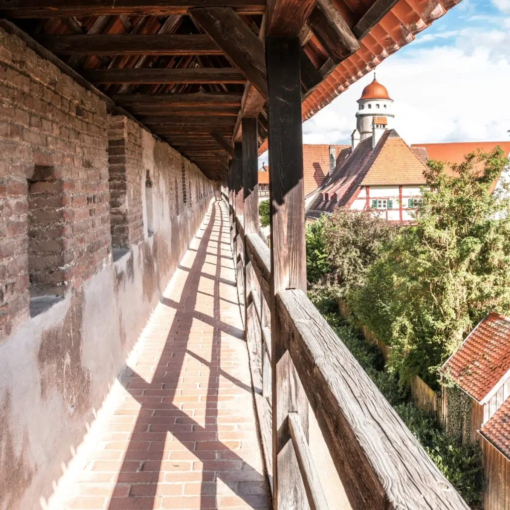Innenansicht des historischen, überdachten Wehrgangs der Nördlinger Stadtmauer mit Holzbalken und Blick auf die Altstadt mit Kirchturm Daniel