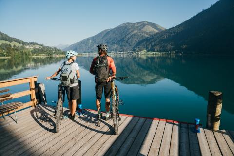Sie sehen zwei Personen mit ihren Mountainbikes am Flussufer des Weissensee.