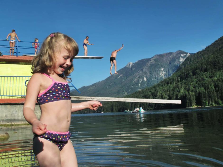 Sie sehen ein Mädchen am Seebad am Lunzer See mit Sprungturm und Menschen im Hintergrund. JUFA Hotels bietet kinderfreundlichen und erlebnisreichen Urlaub für die ganze Familie.