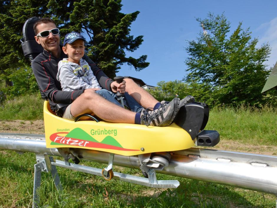 Vater und Sohn sausen mit einer Sommerrodel des Grünberg Flitzers im Salzkammergut ins Tal. JUFA Hotels bietet kinderfreundlichen und erlebnisreichen Urlaub für die ganze Familie.