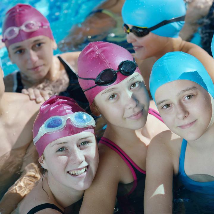 Eine Gruppe von Kindern mit Badehauben stehen im Schwimmbecken und lächeln in die Kamera. JUFA Hotels bietet erholsamen Thermen- und Badespass für die ganze Familie.