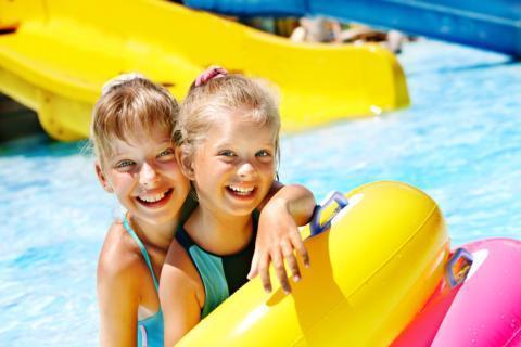 Sie sehen zwei lachende blonde Mädchen mit Schwimmreifen im Freibad mit Rutsche. JUFA Hotels bietet erholsamen Thermen- und Badespass für die ganze Familie.