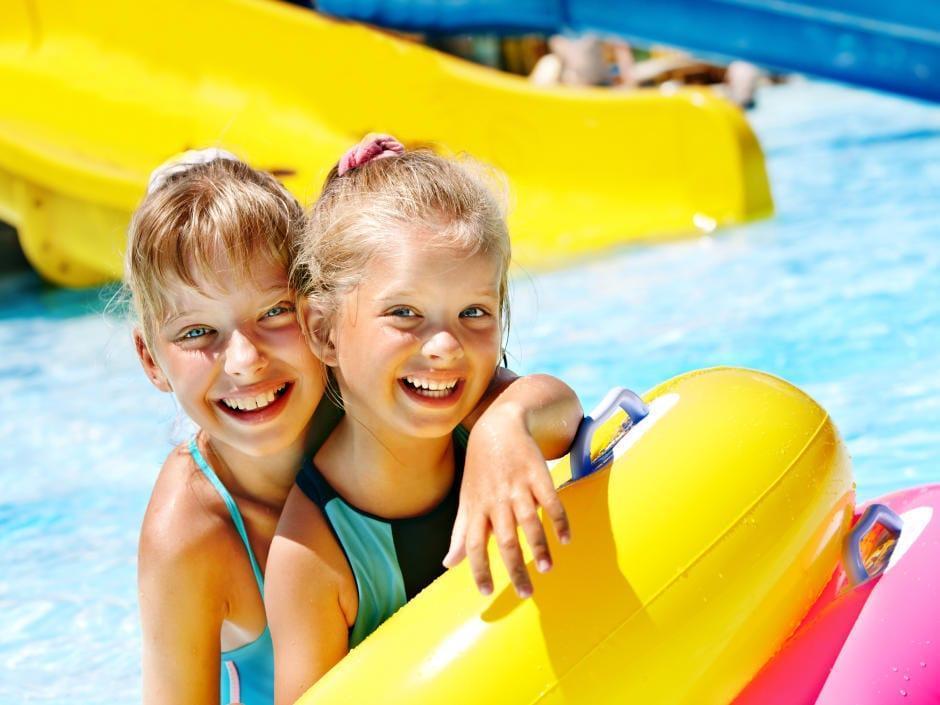 Sie sehen zwei lachende blonde Mädchen mit Schwimmreifen im Freibad mit Rutsche. JUFA Hotels bietet erholsamen Thermen- und Badespass für die ganze Familie.