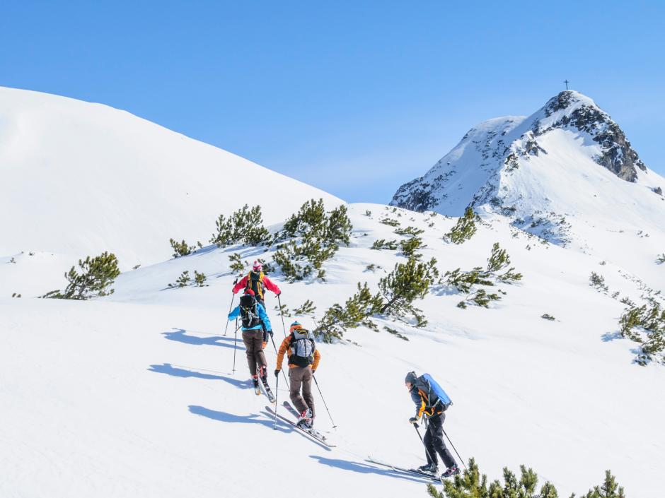 Sie sehen eine Gruppe von 4 Personen, die eine Skitour gehen und im Hintergrund ist der Gipfel mit Gipfelkreuz und eine winterliche Landschaft zu sehen.