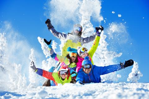 Sie sehen eine Freundesgruppe im Schnee liegend mit Skigewand und Skibrillen umgeben von einer Winterlandschaft.