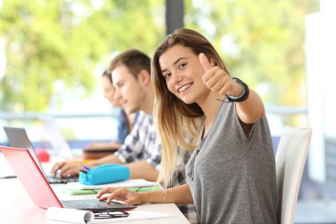 Happy student looking at you with thumbs up in a classroom with classmates in the background