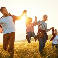 Sie sehen eine Familie mit Mutter, Vater und 2 Kindern, die Hände haltend und mit lächelndem Gesicht über ein Getreidefeld laufen, im Hintergrund ist ein Sonnenuntergang zu sehen.