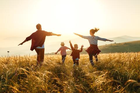 Sie sehen eine Familie mit Mutter, Vater und 2 Kindern, die mit ausgestreckten Armen über ein Getreidefeld laufen, im Hintergrund ist ein Sonnenuntergang zu sehen.