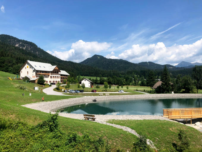Sie sehen das JUFA Hotel Gitschtal Landerlebnis mit Bergsee und Landschaft. JUFA Hotels bietet kinderfreundlichen und erlebnisreichen Urlaub für die ganze Familie.
