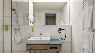 Sie sehen ein modernes Badezimmer mit Waschbecken, Haartrockner, Handtuchtrockner und WC in den Family & Friends Zimmern sowie Suiten im JUFA Hotel Bad Radkersburg.