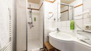Sie sehen ein Badezimmer im JUFA Hotel Seckau mit Dusche und Waschbecken. JUFA Hotels bietet erholsamen Familienurlaub und einen unvergesslichen Winter- und Wanderurlaub.