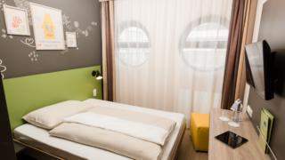 Sie sehen ein Bett im Einzelzimmer mit Fernseher im JUFA Hotel Graz-Süd. Der Ort für erlebnisreichen Städtetrip für die ganze Familie und der ideale Platz für Ihr Seminar.