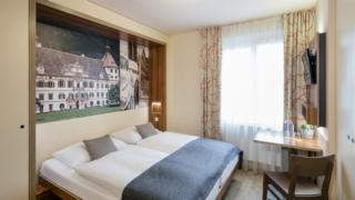 Sie sehen ein Doppelzimmer mit Bett, Kissen und Decke im JUFA Hotel Graz City***. Der Ort für erlebnisreichen Städtetrip für die ganze Familie und der ideale Platz für Ihr Seminar.