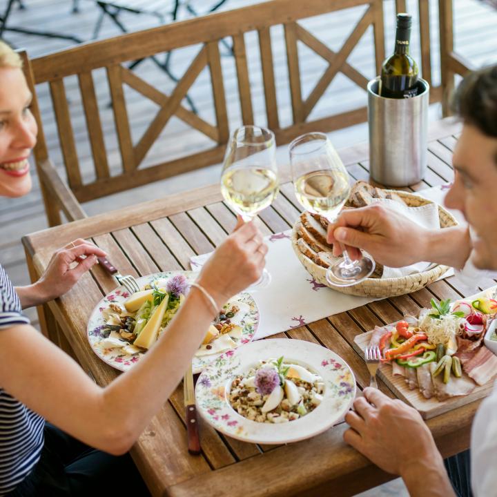 Sie sehen einen Mann und eine Frau an einem gedeckten Tisch mit Köstlichkeiten aus dem Thermen- & Vulkanland beim Anstoßen mit einem guten Glas Wein.