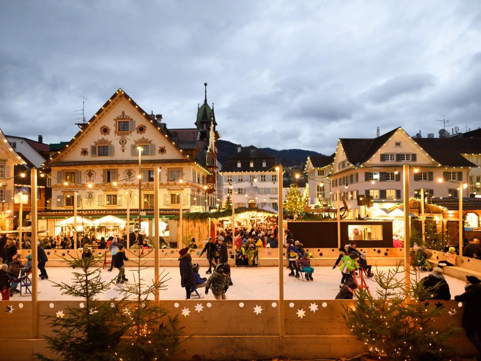 Sie sehen den Christkindlemarkt in Dornbirn in der Abenddämmerung mit festlich geschmückten Häusern und Eislaufplatz
