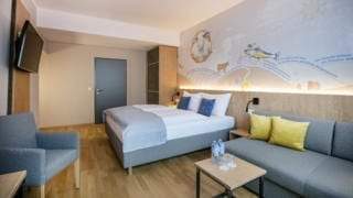 Sie sehen ein Doppelbett und eine Couch im Doppelzimmer im JUFA Hotel Weiz. Der Ort für kinderfreundlichen und erlebnisreichen Urlaub für die ganze Familie.