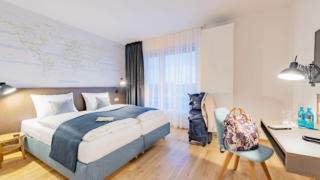Sie sehen ein Doppelbett in einem Doppelzimmer im JUFA Hotel Hamburg HafenCity**** mit einem Tisch. JUFA Hotels bietet erlebnisreichen Städtetrip für die ganze Familie und den idealen Platz für Ihr Seminar.