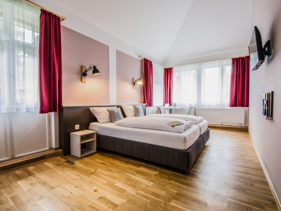 Sie sehen ein Doppelbett in einem Doppelzimmer im JUFA Hotel Königswinter/Bonn mit Fernseher. JUFA Hotels bietet erlebnisreichen Städtetrip für die ganze Familie und den idealen Platz für Ihr Seminar.