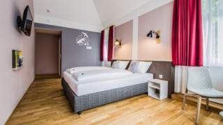 Sie sehen ein Doppelbett in einem Doppelzimmer im JUFA Hotel Königswinter/Bonn mit Stuhl. JUFA Hotels bietet erlebnisreichen Städtetrip für die ganze Familie und den idealen Platz für Ihr Seminar.