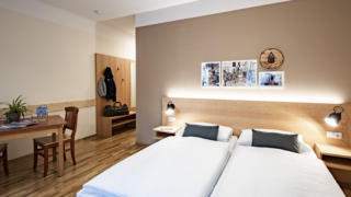 Sie sehen ein Doppelbett in einem Doppelzimmer im JUFA Hotel Murau mit Tisch und Wandbildern. JUFA Hotels bietet erholsamen Familienurlaub und einen unvergesslichen Winter- und Wanderurlaub.