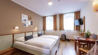 Sie sehen ein Doppelbett in einem Doppelzimmer im JUFA Hotel Murau mit TV und Tisch. JUFA Hotels bietet erholsamen Familienurlaub und einen unvergesslichen Winter- und Wanderurlaub.