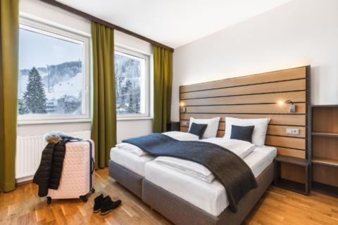 Sie sehen ein Doppelbett in einem Doppelzimmer im JUFA Hotel Schladming mit Koffer. JUFA Hotels bietet erholsamen Familienurlaub und einen unvergesslichen Winterurlaub.