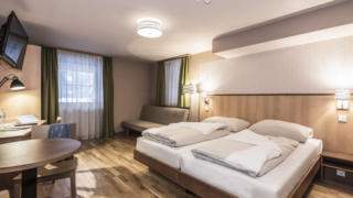Sie sehen ein Doppelbett in einem Familienzimmer medium Plus im JUFA Hotel Grundlsee*** mit einer Sitzgelegenheit. JUFA Hotels bietet erholsamen Familienurlaub und einen unvergesslichen Winter- und Wanderurlaub.