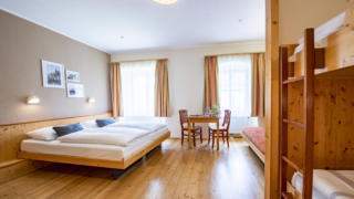 Sie sehen ein Doppelbett in einem Familienzimmer Typ Family and Friends 5 im JUFA Hotel Murau mit Tisch. JUFA Hotels bietet erholsamen Familienurlaub und einen unvergesslichen Winter- und Wanderurlaub.