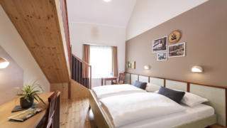 Sie sehen ein Doppelbett in einem Galeriezimmer Typ 6 im JUFA Hotel Murau mit Tisch. JUFA Hotels bietet erholsamen Familienurlaub und einen unvergesslichen Winter- und Wanderurlaub.