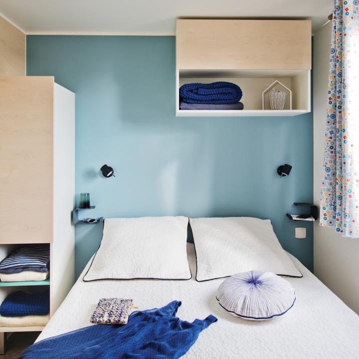 Sie sehen ein Doppelbett in einem Mobile Home beim JUFA Vulkan Thermen-Resort mit großem Fenster. JUFA Hotels bietet erholsamen Thermenspass für die ganze Familie.